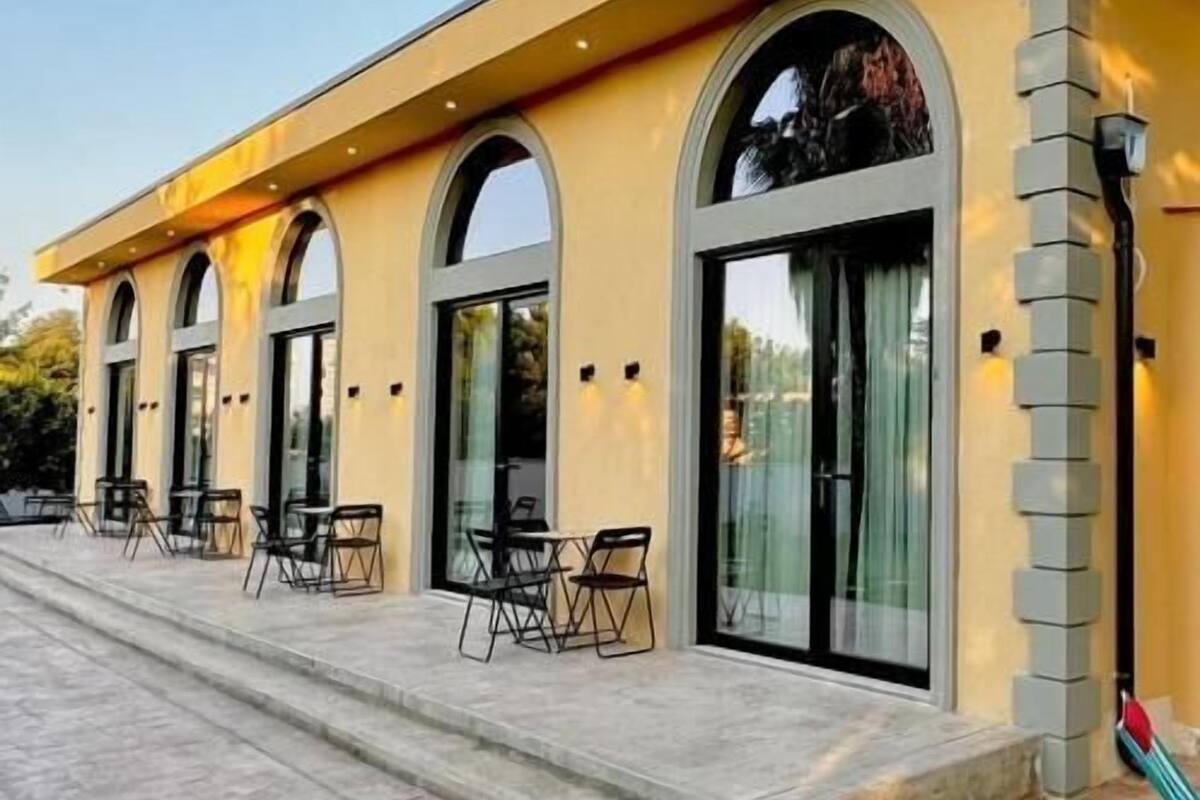 Un edificio con fachada amarilla, grandes ventanales de arco y un área exterior pavimentada con sillas.