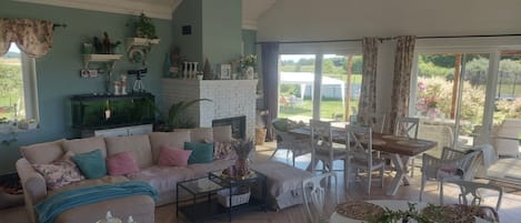 A spacious living room with a fireplace, a dining area with wicker furniture, and a view of the garden through large sliding doors.