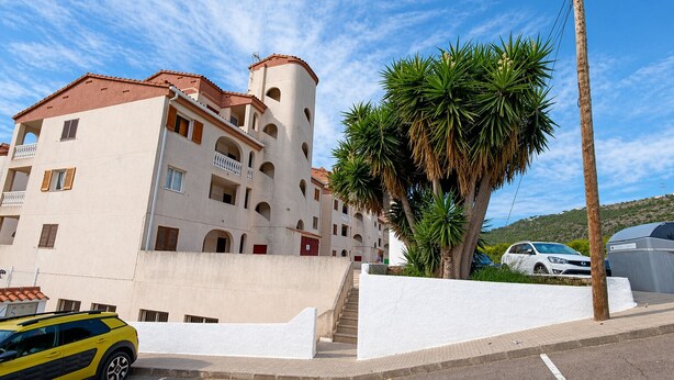 A multi-story building with balconies, a staircase leading to the entrance, and a parking area with a yellow car. - Charming Alcocéber Apartment With Terrace and Pool Access (Alcala de Xivert)