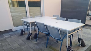 An outdoor patio with a white table and blue chairs, surrounded by a tiled floor and a dark wall. - Ferienwohnung in Petersdorf auf Fehmarn (Petersdorf)