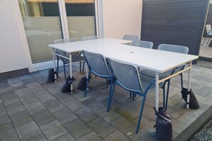 Terrassa exterior amb taula blanca i cadires blaves, envoltada per un paviment de rajoles i una paret fosca.