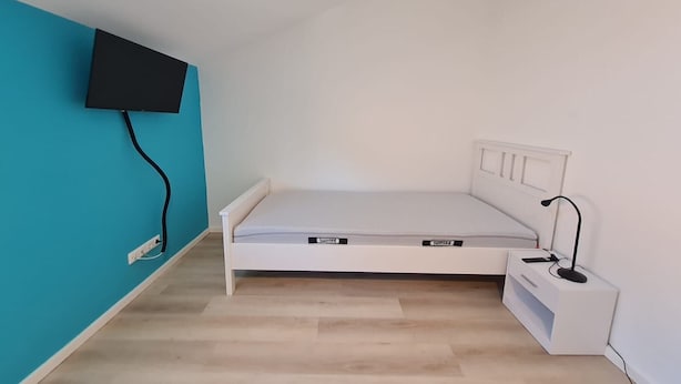 A small, single-bed room with a white bed, a bedside table, a lamp, and a TV mounted on the wall. - Ferienwohnung in Petersdorf auf Fehmarn (Petersdorf)