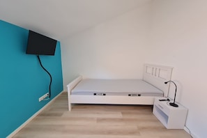 A small, single-bed room with a white bed, a bedside table, a lamp, and a TV mounted on the wall. - Ferienwohnung in Petersdorf auf Fehmarn (Petersdorf)
