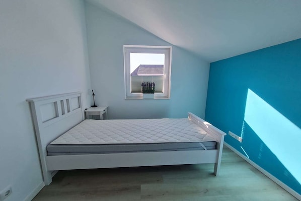 Ein Schlafzimmer mit einem Bett, einem kleinen Nachttisch, einem Fenster und einer blauen Wand.