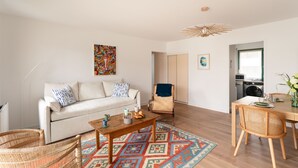 Ein Wohnzimmer mit einer Couch, Couchtisch und Sesseln, ein Essbereich mit Tisch und Stühlen sowie eine Küche im Hintergrund.