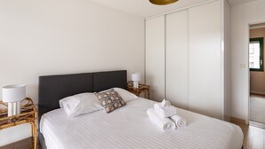 Ein ordentlich bezogenes Bett mit weißen Leinen, ein dunkles Kopfstück und ein dekoratives Kissen. Ein kleiner Nachttisch mit Lampe und ein Korbstuhl sind vorhanden. Im Hintergrund ist ein großer, weißer Kleiderschrank zu sehen.