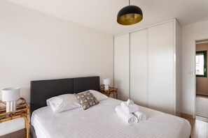 Una cama bien hecha con sábanas blancas, un cabecero oscuro y un cojín decorativo. Hay una mesita de noche con una lámpara y una silla de mimbre. Se observa un amplio armario blanco al fondo.