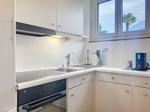 A kitchen with a sink, stove, cabinets, and a window with a view of palm trees. - Residenza Corallo by Interhome (Ascona)