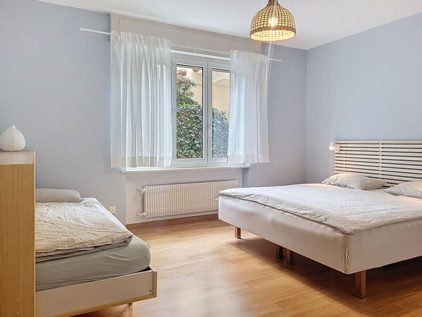 A bedroom with two beds, a window with curtains, a radiator, and a hanging light fixture. - Residenza Corallo by Interhome (Ascona)