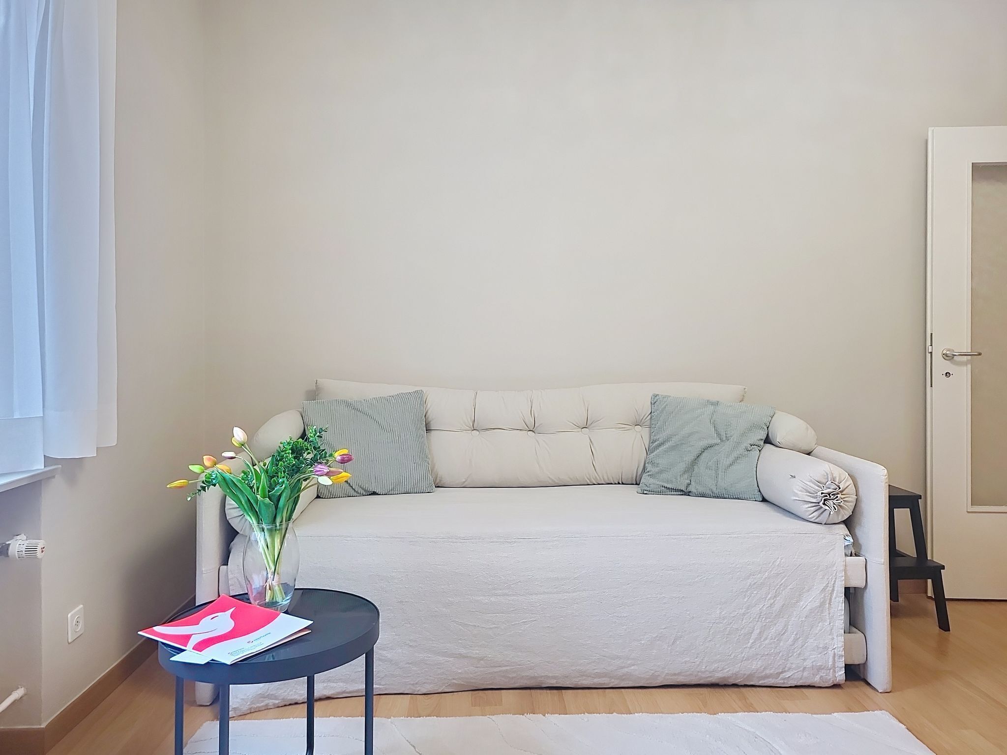 Un canapé de couleur neutre agrémenté de deux coussins, une petite table ronde sur laquelle est posé un vase de fleurs, et un tapis blanc sur un plancher en bois.