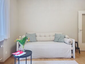 Ein sofasitz in neutralen Farben mit zwei Kissen, ein kleiner runder Tisch mit Blumen in einer Vase und ein weißer Teppich auf Holzdielen.