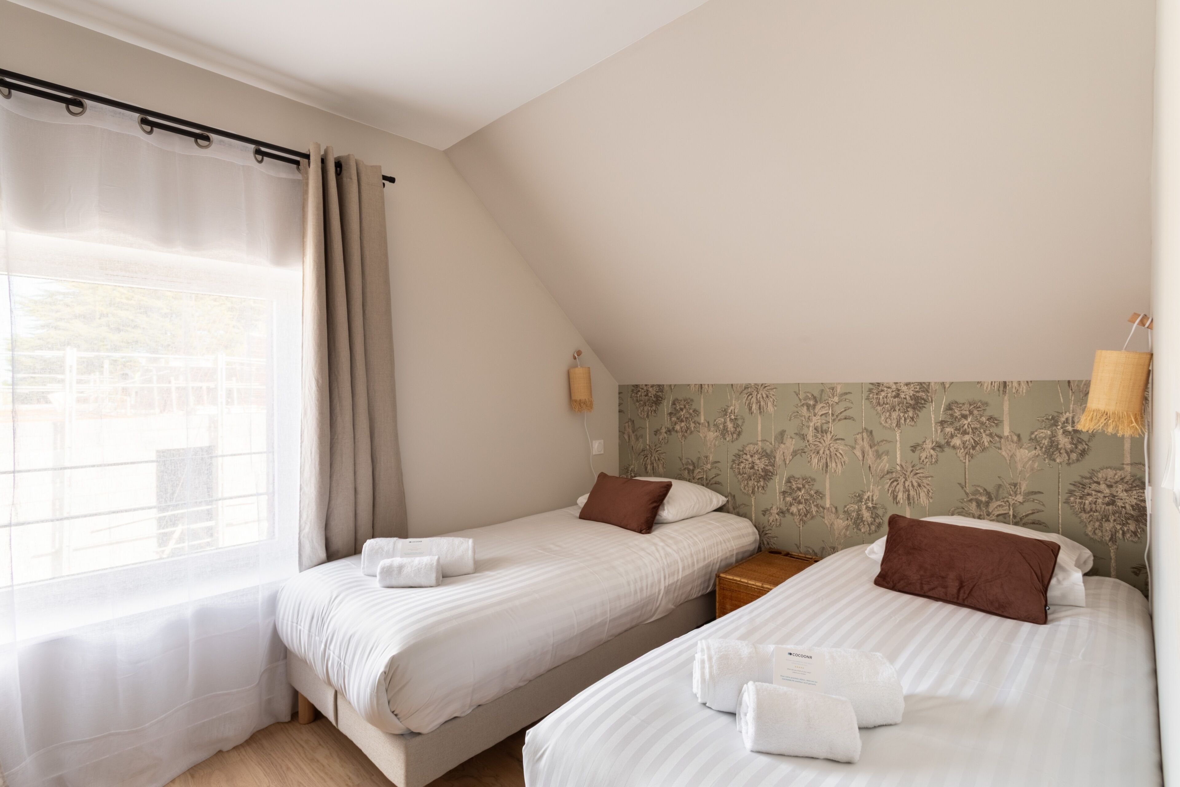 A bedroom with two beds, a window with curtains, a nightstand, and a wall-mounted lamp.