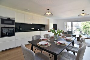 A modern kitchen with white cabinets, a dining table set with plates and glasses, and a large window with a view. - Résidence Des Champs sur Mer - À 400 m de la plage (Dinard)