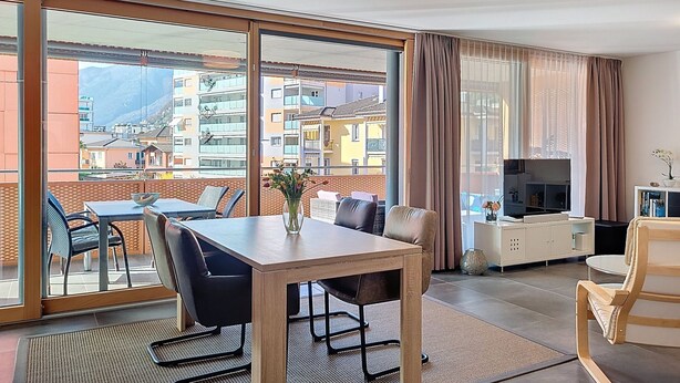 A modern hotel room with a dining table, chairs, a sofa, a TV, and large windows offering a city view. - Residenza Eride - Apt.4 by Interhome (Locarno)