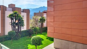 A modern building with a red facade, a well-maintained garden with a small tree, and a view of distant mountains. - Residenza Eride - Apt.4 by Interhome (Locarno)