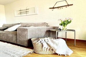 A cozy living room with a sofa, a small round table with a vase of flowers, and a decorative wall piece. - Ferienwohnung in Lemkenhafen für 3 Gäste (Lemkenhafen)