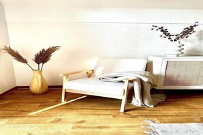 A room with a wooden floor, a white chair, a wooden side table, a white radiator, and a vase with dried plants. - Ferienwohnung in Lemkenhafen für 3 Gäste (Lemkenhafen)
