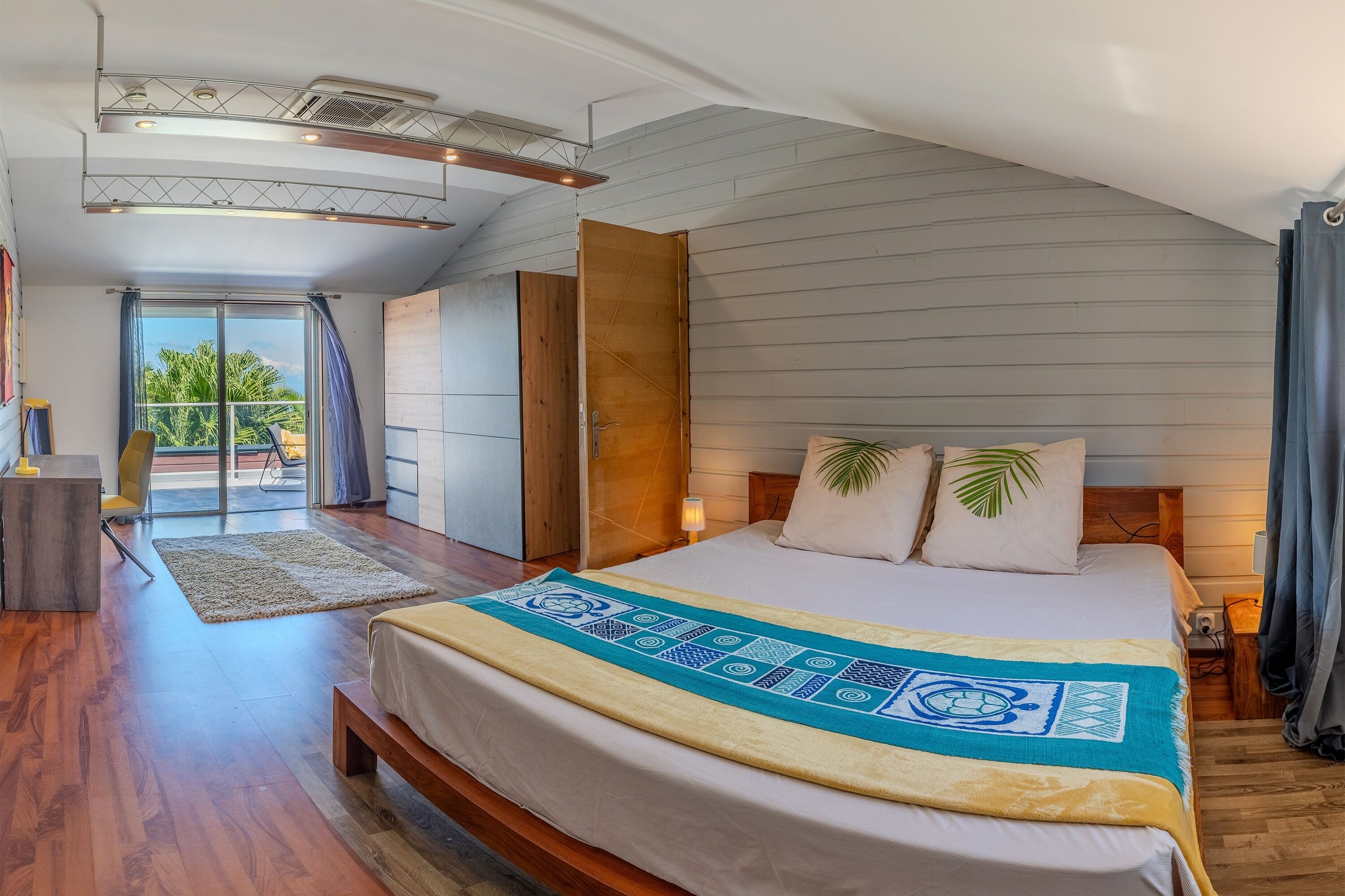 A bedroom with a bed, a wooden bench, a nightstand, a lamp, a window with curtains, and a view of the outdoors.