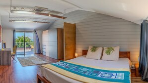 Ein Schlafzimmer mit einem Bett, einer Holzbank, einem Nachttisch, einer Lampe, einem Fenster mit Vorhängen und Blick ins Freie.