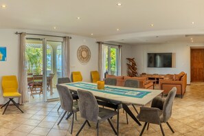 A spacious dining area with a long table, multiple chairs, and a view through glass doors to an outdoor patio. - La Case des Fleurs de Jade - Avec piscine-jacuzzi (Petite Île)