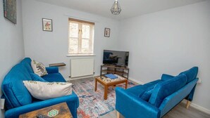 Un salon avec deux canapés bleus, une table basse, un téléviseur posé sur un meuble et une fenêtre avec des stores.