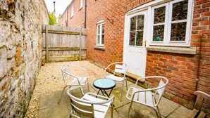 Un bâtiment en briques avec une porte et des fenêtres blanches, une clôture en bois et une terrasse avec des chaises et une table en métal.