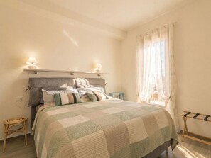 A bedroom with a bed, bedside lamps, a window with curtains, and a small stool. - Nuraghi by Interhome (Arzachena)