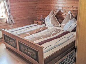 A wooden bed with a patterned bedspread and pillows, a nightstand with a lamp, and a window with curtains.