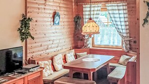 Interior acolhedor de uma cabana, com parede de madeira, um espaço de estar com bancos, uma mesa e uma televisão.