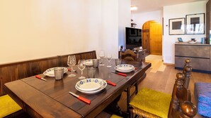 Uma área de refeições com uma mesa de madeira posta para quatro pessoas, uma televisão ao fundo e uma cozinha com um armário de madeira e uma pia.