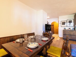 Ein Essbereich mit einem Holztisch, gedeckt für vier Personen, einem Fernseher im Hintergrund und einer Küchenzeile mit Holzschrank und Spüle.