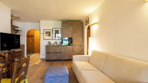 Uma sala de estar com sofá, apoio para os pés, mesa de jantar de madeira, cadeiras, televisão e área de cozinha com armários e eletrodomésticos.