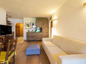 Ein Wohnzimmer mit einer Couch, Hocker, einem Holzessttisch, Stühlen, einem Fernseher und einer Küchenzeile mit Schränken und Geräten.