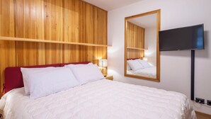 Quarto de hotel com parede de painel de madeira, cama com roupa de cama branca, cabeceira vermelha, mesa de cabeceira com luminária e televisão fixada na parede.