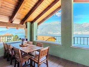 Ein Holztisch mit Stühlen, eine Vase mit Blumen und eine Obstschale auf einem Balkon mit Blick auf Berge und einen See.