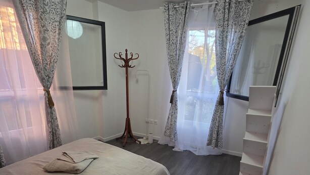 A bedroom with a bed, a coat rack, a window with curtains, a radiator, and a staircase. - Escale Zen & Loggia: 5 min from the train station (Direct Paris) + Parking (Évry-Courcouronnes)