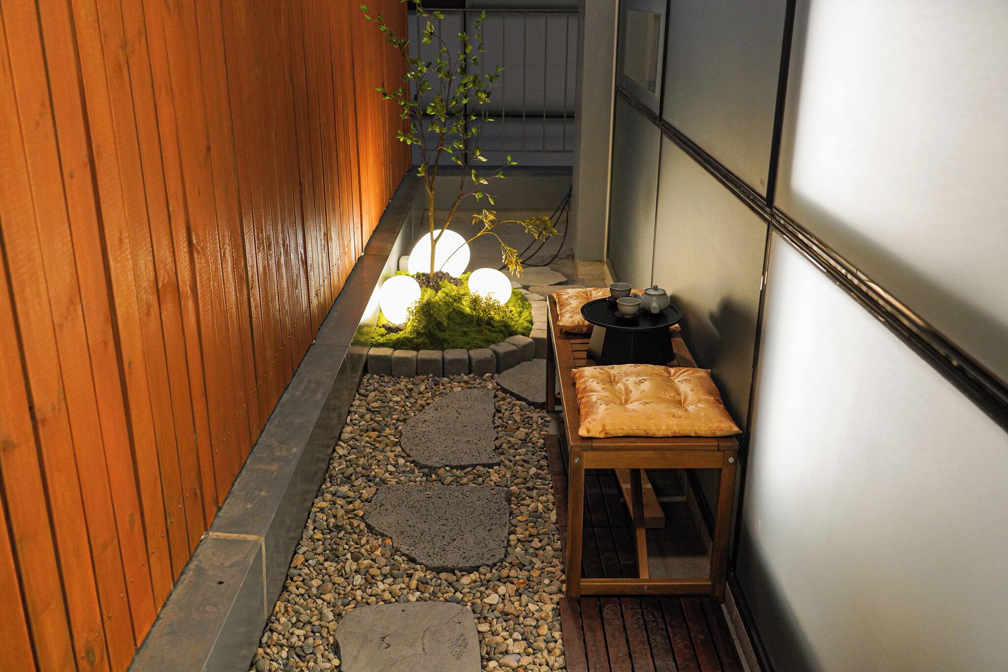 Un estrecho sendero con piedras para pisar, un banco de madera, una pequeña mesa con tazas, y una planta cubierta de musgo.
