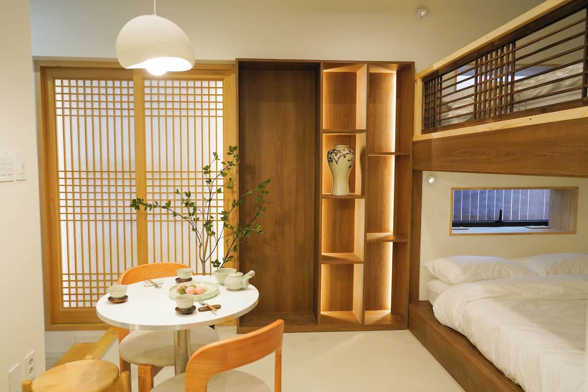 Una habitación con una cama litera, una mesa de comedor con sillas, un estante pequeño y una ventana.