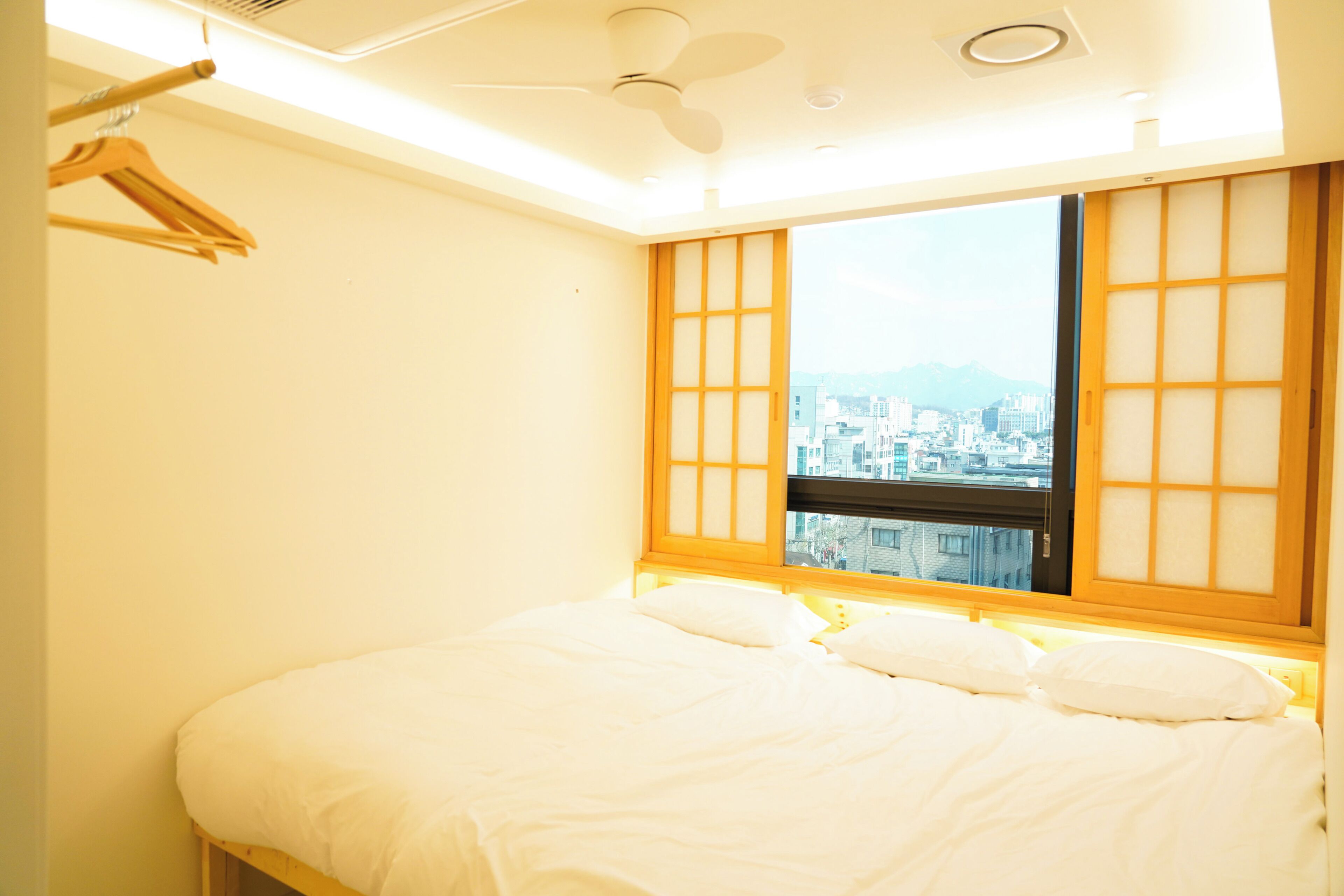 A bedroom with a bed, a window with a view of a cityscape, a ceiling fan, and a wooden wardrobe.