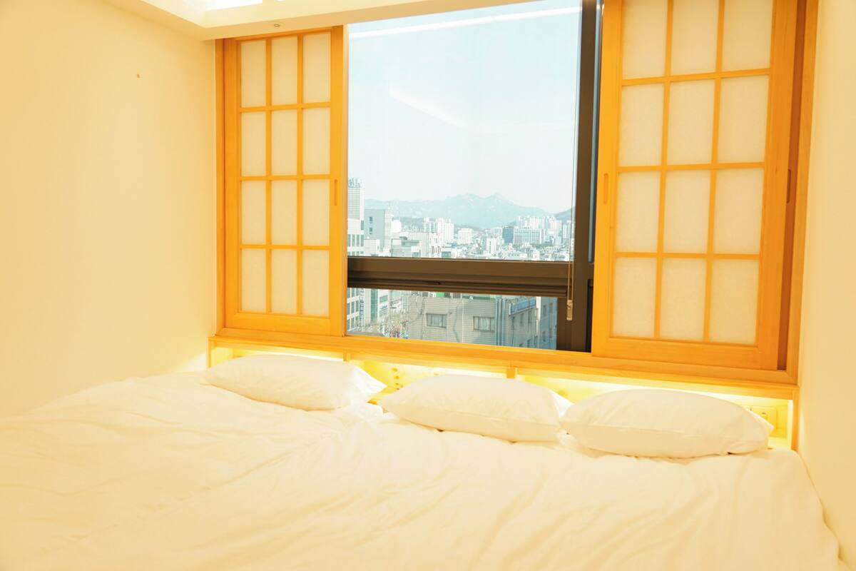 Una habitación con una cama, una ventana con marcos de madera y vistas a un paisaje urbano.