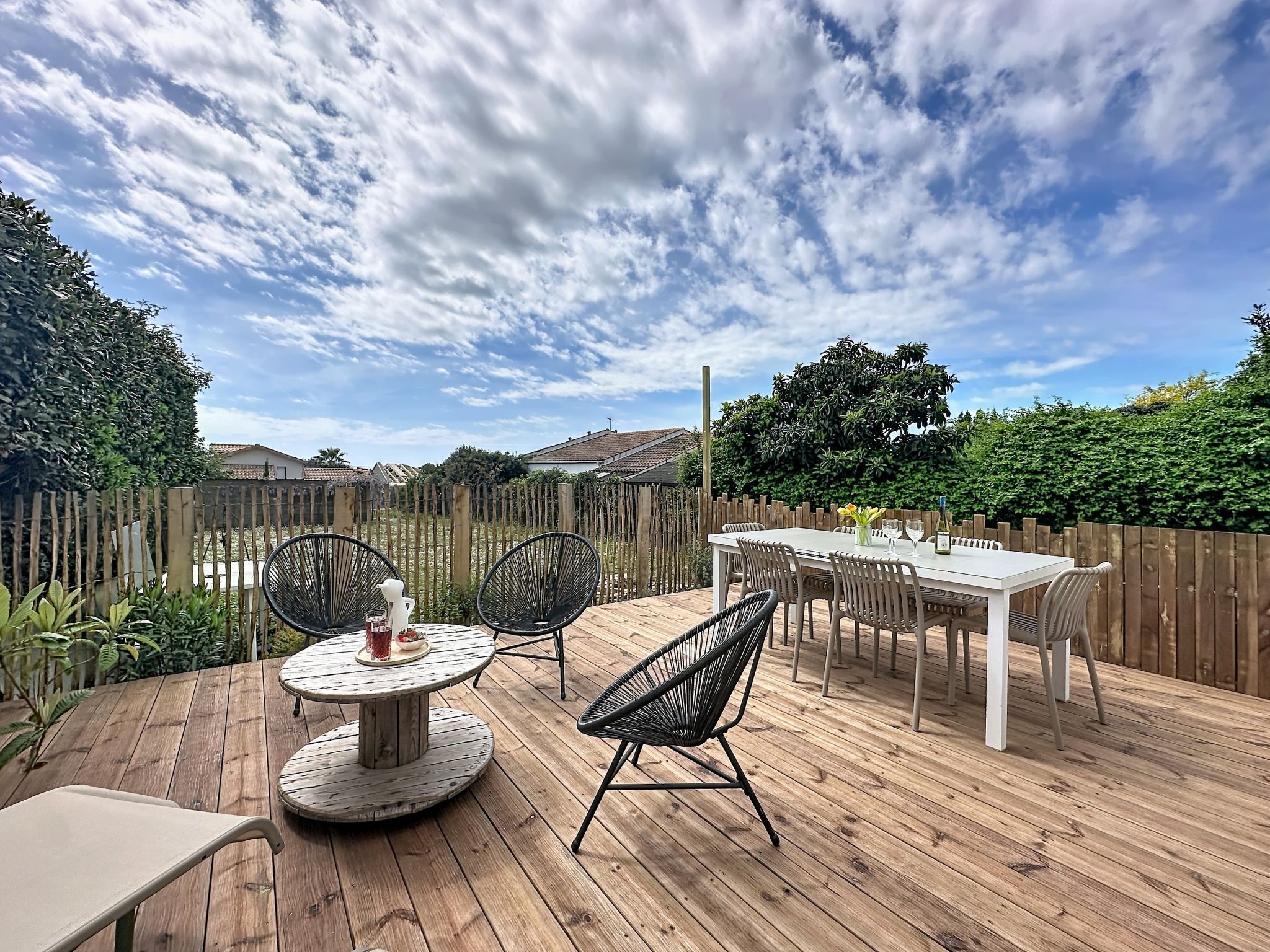 A wooden deck with a round table and chairs, surrounded by a wooden fence and greenery.