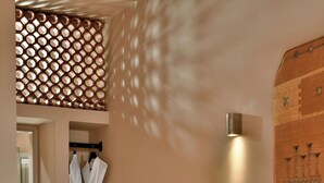 A bedroom with a bed, a hanging robe, a wall-mounted light, and a decorative wall panel. - Terra Verde Oia Suites - Sienna Premium Room (Oia)
