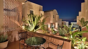 A terrace with a round table and chairs, surrounded by potted plants and a decorative lattice wall. - Terra Verde Oia Suites - Sienna Premium Room (Oia)