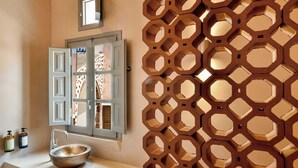 A bathroom with a sink, a window with open shutters, and a decorative wall panel. - Terra Verde Oia Suites - Sienna Premium Room (Oia)