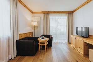 Una habitación con piso de madera, un sofá, una pequeña mesa redonda con una lámpara, un televisor sobre un mueble de madera y una ventana con cortinas.