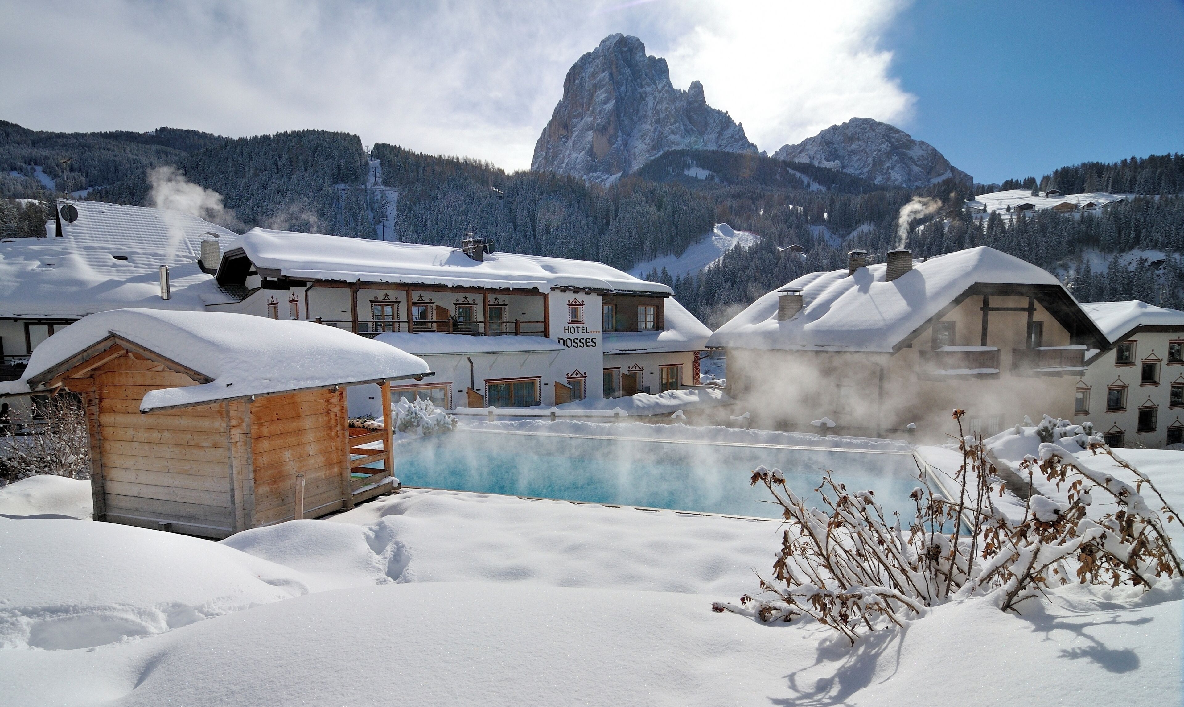 A hotel with a pool surrounded by snow, wooden cabins, and mountains in the background.