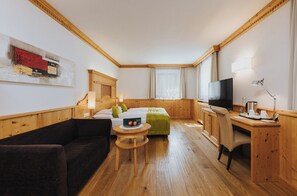 A hotel room with a wooden floor, a bed, a desk with a chair, a television, and a small round table with apples. -  Vitalhotel Dosses (Santa Cristina Val Gardena)