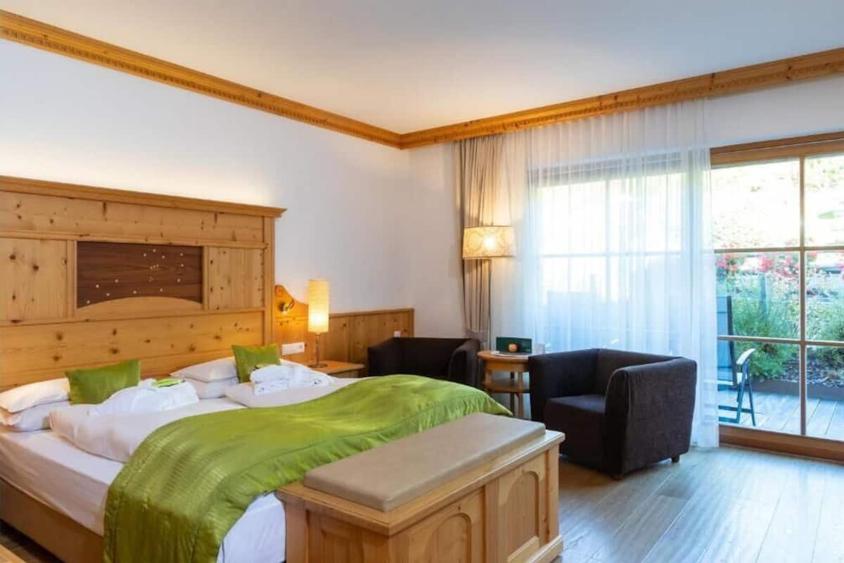 A hotel room with a wooden headboard, a bed with green bedding, a bedside table with a lamp, a chair, and a view of the outdoors through large windows.