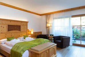 Habitación de hotel con cabecera de madera, cama con ropa de cama verde, mesita de noche con lámpara, una silla y vistas al exterior a través de amplias ventanas.