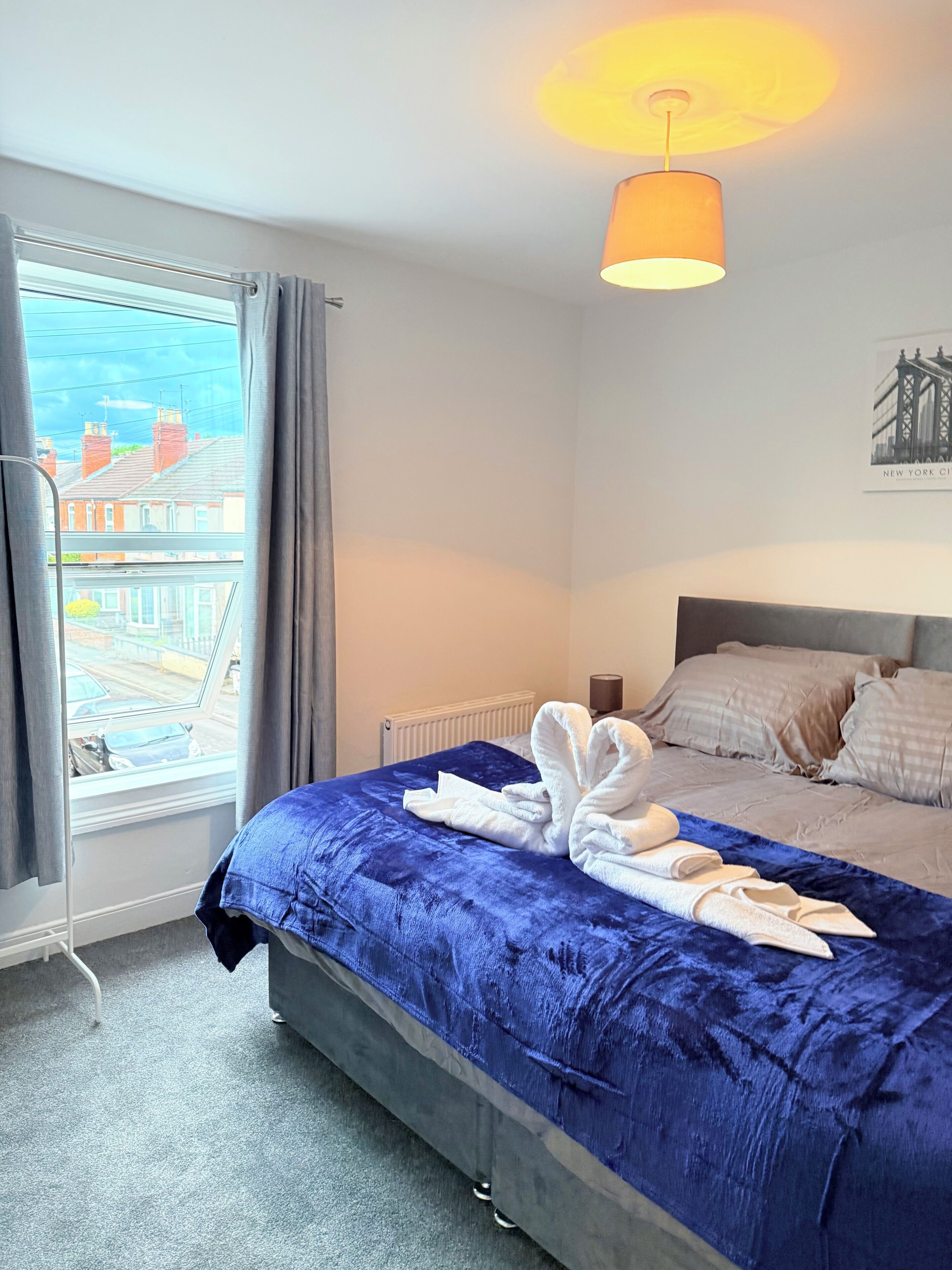 A hotel room with a large bed, a view of the city through a window, a radiator, and a ceiling light.